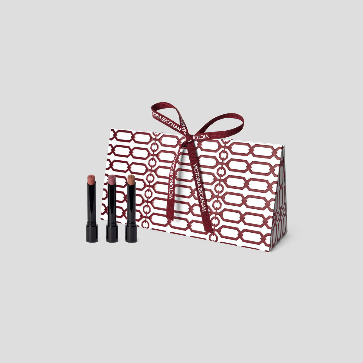 Three Posh Lip lipsticks in front of a burgundy trellis gift box with branded bow — Victoria Beckham Beauty Posh Lip trio.