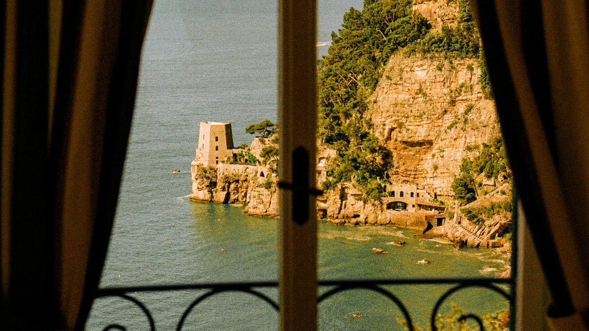 Stone tower and pine-covered cliffs above a green sea, through a curtained window — Hotel Portofino by Victoria Beckham Beauty.
